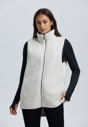 Fleece vest in off-wit met hoge kraag, volledige rits en zijzakken; gelaagd over een zwart T-shirt met lange mouwen, met een zachte textuur.
