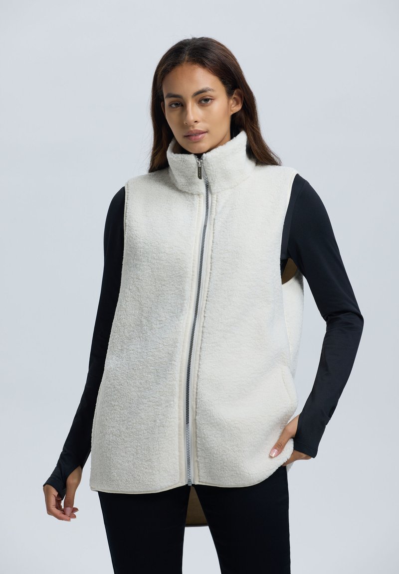 Fleece vest in off-wit met hoge kraag, volledige rits en zijzakken; gelaagd over een zwart T-shirt met lange mouwen, met een zachte textuur.