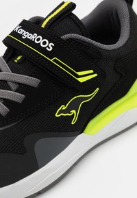 Schwarzer Sportschuh mit einem grauen Mesh-Obermaterial, gelben Akzenten, einem Klettverschluss und einem Känguru-Logo. Verfügt über eine weiße Sohle mit einem strukturierten Design.