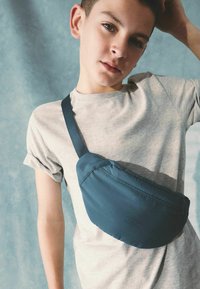 Banane bleu marine en tissu doux, de forme incurvée avec une sangle ajustable, portée sur un t-shirt gris clair. Design simple et fonctionnel.