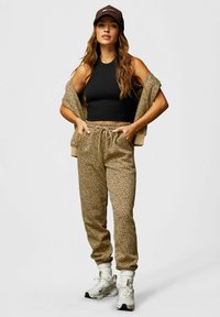 Frau in einem schwarzen ärmellosen Crop-Top, beigen gepunkteten Jogginghosen, passender Jacke über den Schultern, beiger Cap, weißen Socken und Sneakers, die mit den Händen in den Taschen steht.