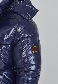 Navy blue puffer jacket with glossy texture, featuring a high collar, snap buttons, and a gold-accented logo patch on the sleeve.