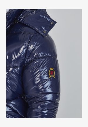 Marineblå puffjakke med blank tekstur, der har høj krave, trykknapper og et logo-patch med guldaccenter på ærmet.