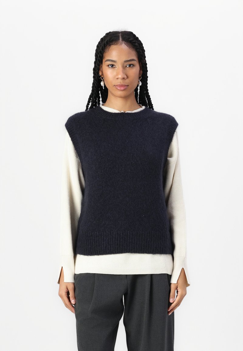 Sweater sem mangas de lã na cor azul-marinho, com um acabamento texturizado, usado sobre uma blusa de manga longa cremosa. Apresenta decote redondo e bainha canelada.
