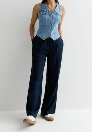 Femme portant un gilet en jean bleu sans manches boutonné, un pantalon ample bleu foncé, des baskets blanches, debout les mains dans les poches devant un fond uni.
