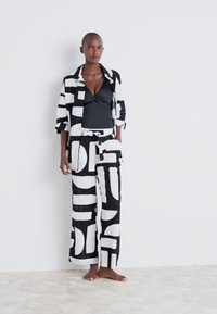 Ensemble v beli in črni barvi z vzorcem, ki vključuje krajši top in široke hlače, izdelan iz lahkega materiala. Oblikovni elementi v abstraktnih oblikah prevladujejo v designu.