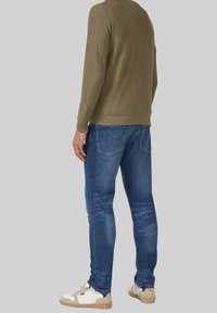 Mężczyzna w oliwkowozielonej koszuli z długim rękawem, niebieskich dżinsach i białych trampkach stoi na tle jednolitego szarego tła.