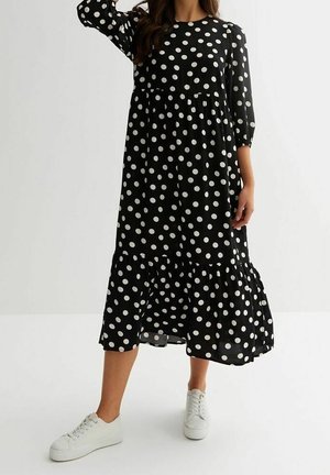 Woman wearing a black midi dress with white polka dots and ruffled hem, paired with white sneakers, standing against a plain background.