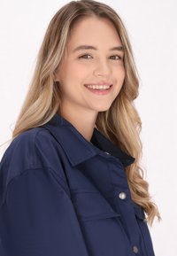 Námořnický modrý sako na knoflíky s velkým límcem, obsahující dvě přední kapsy a hladký povrch. Modelka má dlouhé, vlnité vlasy.