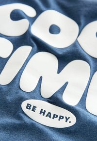 Blåt stof med hvidt fed tekst, der siger "COOL" og "BE HAPPY." Teksten har rundede former, en glat tekstur og en let glans.