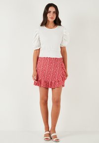 Witte textuur top met pofmouwen, gecombineerd met een rode bloemenrokken met ruffles. Sandalen hebben een gevlochten bandontwerp.