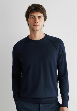 Ung mand med kort krøllet brunt hår iført en tætsiddende marineblå langærmet rundhals-sweater, stående mod en ensfarvet lys baggrund.