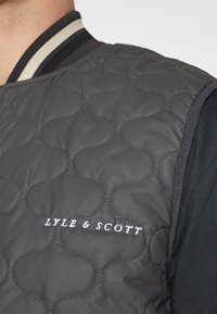 Quilted gray vest with black trim and striped collar. Features "LYLE & SCOTT" embroidered in white on the front. Textured pattern visible.