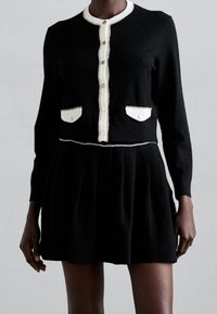 Svart långärmad kort cardigan med vit knapprand och fickor, tillsammans med en plisserad svart kjol på en stående person.