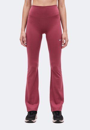 Nő magas derekú, rózsaszín színű, bővülő szárú leggings-ben, bal combján kicsi fehér Nike logóval, fekete-fehér sportcipővel párosítva.