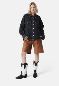 Cămașă din denim, albastră închisă, cu nasturi și buzunare pe piept; pantaloni scurți din piele ecologică maro cu textură; cizme în stil vestic alb-negru cu vârfuri ascuțite.