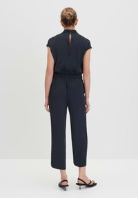 Marine jumpsuit met korte mouwen, elastische taille en een sleufopening op de rug; het materiaal lijkt glad. Gecombineerd met zwarte hakken sandaaltjes.