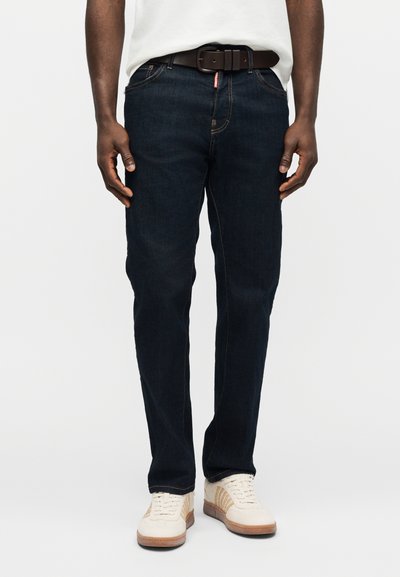 Άνδρας που φοράει σκούρα μπλε τζιν, λευκά αθλητικά παπούτσια, καφέ ζώνη και λευκό πουκάμισο μπαίνωντας μέσα στο παντελόνι, στέκεται με τα χέρια χαλαρά δίπλα στο σώμα.