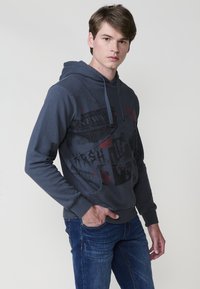 Felpa navy blue con stampa grafica che presenta testi e immagini correlati alle notizie. Materiale in misto cotone, con cappuccio regolabile e tasca frontale.