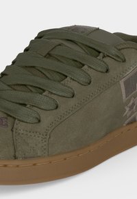 Gros plan d'une basket en daim vert olive avec des lacets assortis et une semelle en caoutchouc marron, présentant des détails cousus et un patch logo latéral.
