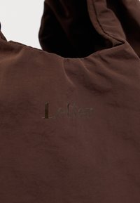 Καφέ ύφασμα τσάντα με μαζεμένο πάνω μέρος και μαύρο λογότυπο "LeGer" τυπωμένο στο μπροστινό μέρος.