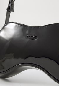 Schwarze Handtasche aus lackiertem Leder mit wellenförmigem Design, das eine strukturierte Oberfläche und ein Logodetail aufweist. Einzelner verstellbarer Träger.