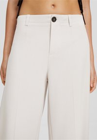 Ljusbeiga vida byxor gjorda av ett slätt tyg. Har en framknäppning och sidofickor med en ren, skräddarsydd design.