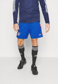 Muž oblečený v modrých šortkách Bayern Munich, námořnickém dlouhém rukávu, černých ponožkách Adidas a černých kopačkách stojící na bílém pozadí.