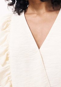 Ljus beige texturerad plagg med volangdetaljer längs axeln och en djup V-urringning, som ger ett mjukt och flödande utseende.