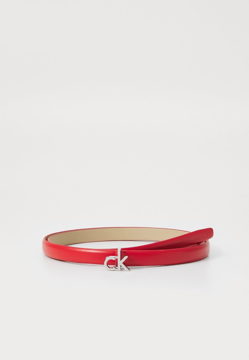 Ceinture fine en cuir rouge roulée avec une boucle argentée du logo CK sur fond blanc.