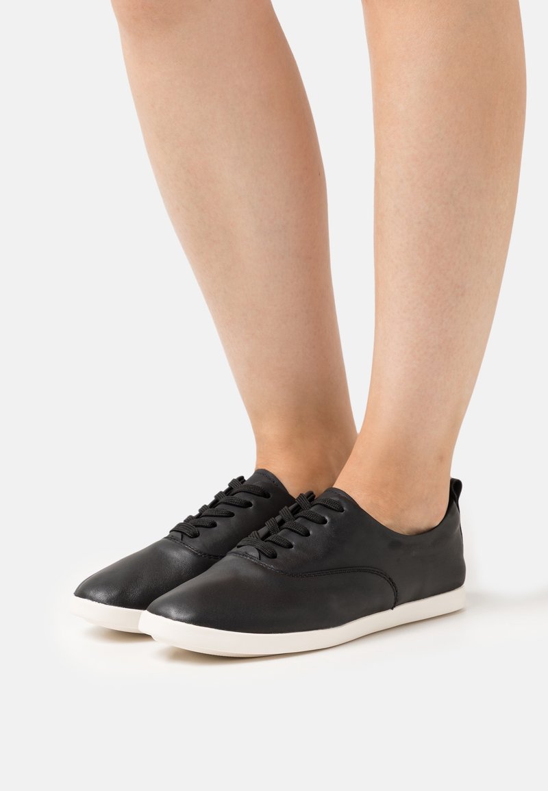 Personne portant des baskets basses noires avec des semelles blanches, debout sur une surface blanche unie.