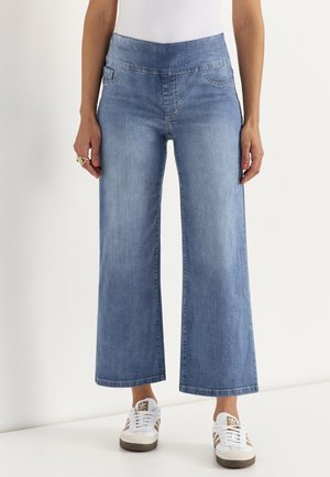 Nainen, jolla on vaaleansiniset leveälahkeiset polvipituiset farkut, joissa on joustava vyötärönauha, ja valkoiset lenkkarit, joissa on ruskeat raidat, seisoo valkoista taustaa vasten.