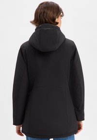 Veste softshell noire avec une capuche ajustée, une texture lisse et des coutures latérales réglables. Elle présente un logo sur le col et une coupe mi-longue.