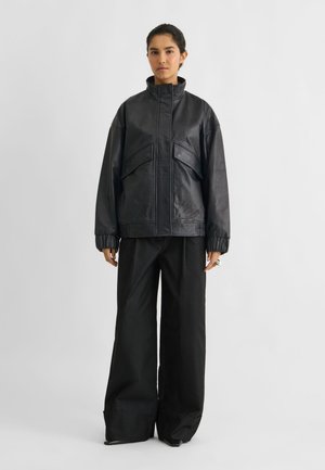 Un mannequin se tient devant un fond de studio clair, vêtu d’une veste en cuir noire, d’un pantalon à jambes larges et de chaussures noires.