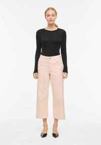 Femme portant un haut noir moulant à manches longues, un pantalon large rose clair coupé au-dessus de la cheville, et des talons pointus noirs, debout devant un fond blanc.