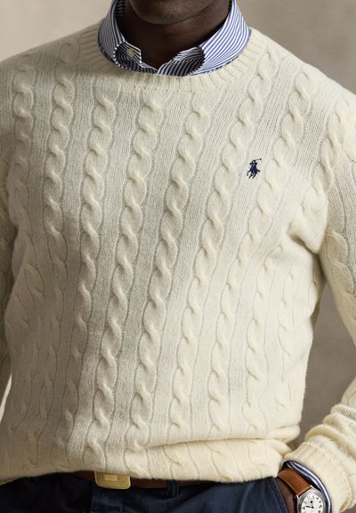 Mand iført en cremefarvet kabelstrikket sweater med blåt broderet logo over en blå- og hvidstribet skjorte og mørke bukser med brun bælte.