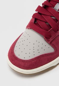 Bordo tenisica od brušene kože sa sivim prstnim poklopcem od kože, perforiranim detaljima i crvenim vezicama. Ima bijelu srednju potplatu i bež gumeni vanjski potplat.