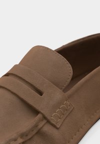 Ανοιχτό καφέ μοκασίνι από σουέτ με ραμμένη λεπτομέρεια λουριού και λείος στρογγυλεμένος μύτη, φωτογραφημένο σε απλό λευκό φόντο.