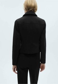 Veste noire courte au design ajusté avec une texture subtile. Elle présente un large col et une silhouette arrière épurée.