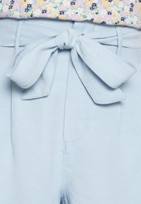 Světle modré kalhoty s uvázanou mašlí v pase. Mají plochý přední díl, boční kapsy a hladký textil.