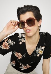 Óculos de sol oversized castanhos com uma armação grossa e translúcida, combinados com um top preto com padrão de flores, apresentando detalhes em rosa e amarelo.