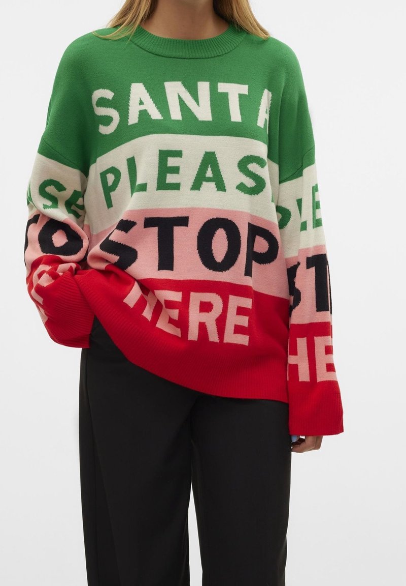 Femme portant un pull multicolore avec le texte "Santa Please Stop Here" en blocs verts, blancs, roses et rouges, assorti à un pantalon noir.