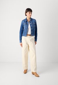 Giubbotto in denim di blu medio, con design classico e tasche frontali, abbinato a pantaloni a gamba larga color avorio e scarpe con tacco color beige.