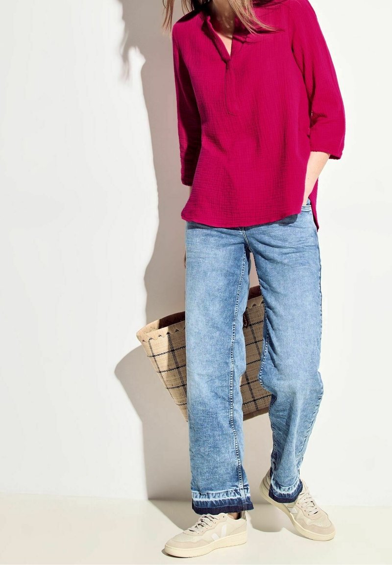 Haut en coton cramoisi avec col, manches trois-quarts ; associé à un jean en denim bleu clair à coupe décontractée ; baskets blanc cassé ; sac fourre-tout en paille tressée.