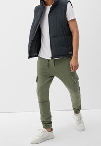Dunkelblau gepolsterte Weste mit hohem Kragen über einem weißen T-Shirt, kombiniert mit olivgrünen Jogginghosen mit Taschen und elastischen Bündchen. Weiße Sneakers.