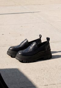 Par sorte plateau loafers med chunky såler og sølv nitter, placeret på en lys beton gulv i naturligt sollys.