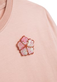 Rosa Sweatshirt mit einem floralen Brosche. Die Brosche besteht aus sich überlappenden Pailletten in Rosa und Weiß, die von einem braunen Faden umrandet sind.