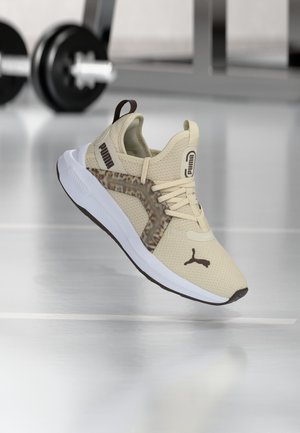 Pantof sportiv Puma bej cu logo maro şi suport lateral cu model, plutind deasupra unei săli de gimnastică cu greutăţi neclare în fundal.