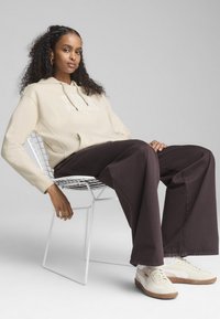 Beige hoodie med "sports club" tekst, parret med mørkebrune brede bukser og cremefarvede sneakers, siddende på en hvid trådstol.