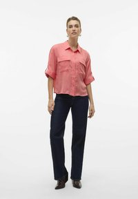 Camicia button-up rosa con due tasche sul petto, maniche arrotolate, abbinata a jeans dritti in denim scuro e stivaletti con tacco marroni.